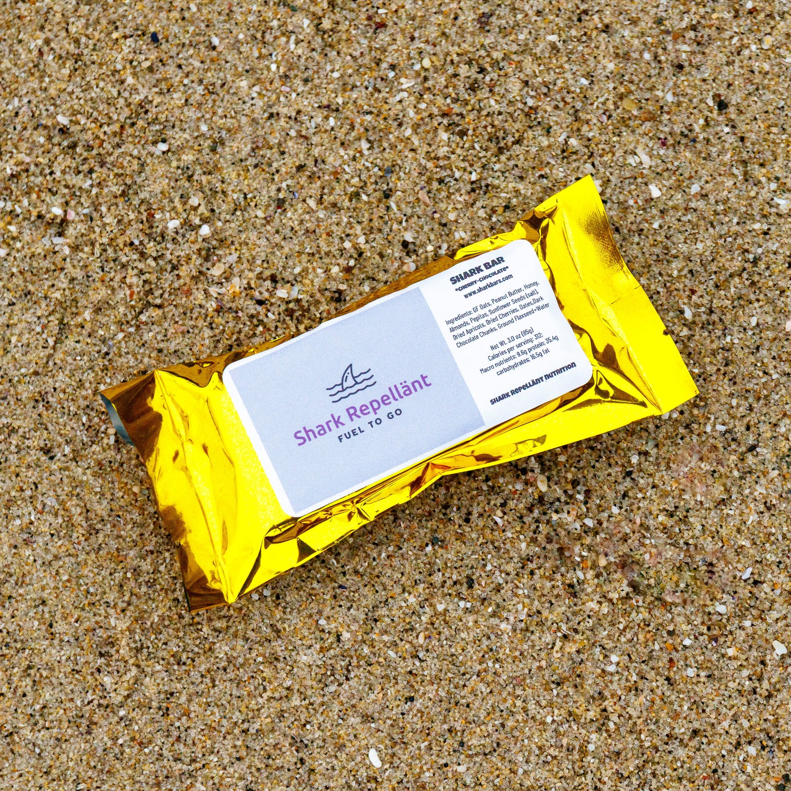 Gold foil wrapper with label of a Cherry Chocolate Gluten Free Oat Honey Shark Bar laying on well packed beach sand
