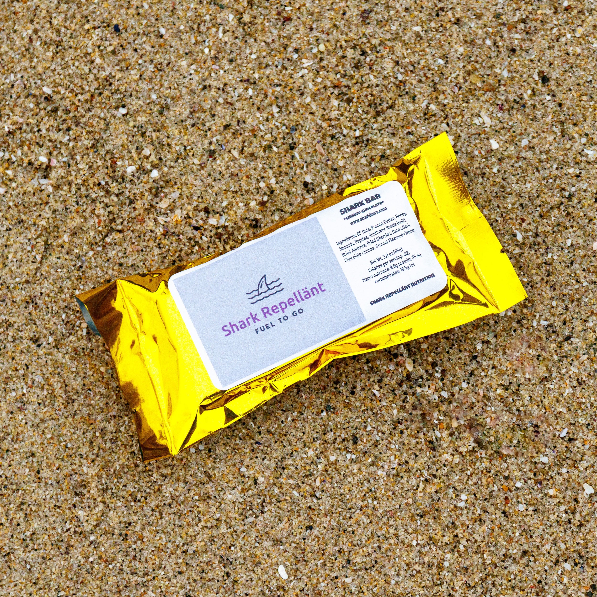Gold foil wrapper with label of a Cherry Chocolate Gluten Free Oat Honey Shark Bar laying on well packed beach sand