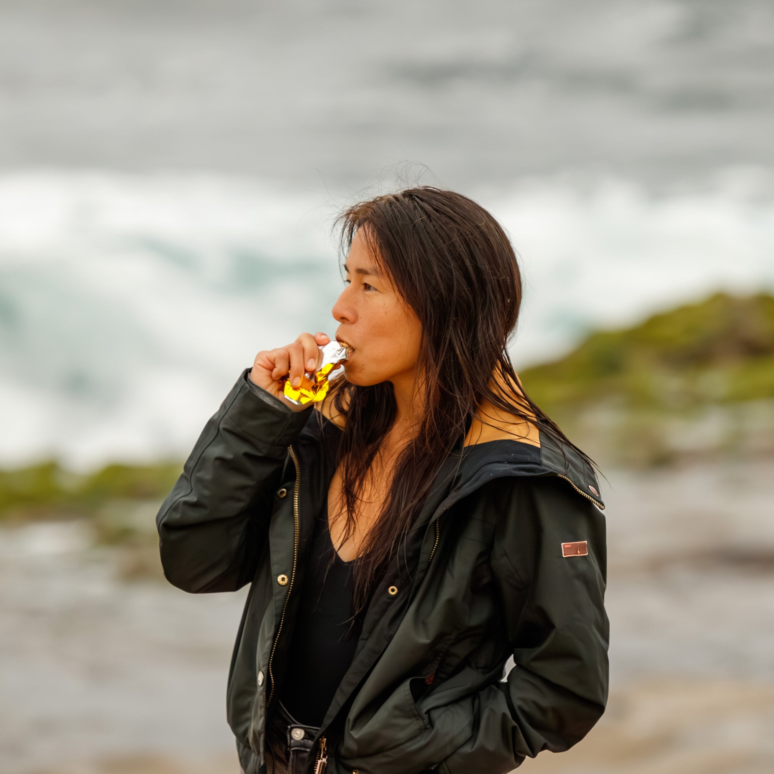 Woman in a black jacket eating a gold foil wrapped cherry chocolate Shark Bar with gluten free oats and honey standing in front of a blurred natural ocean cliff background along the southern california coastline