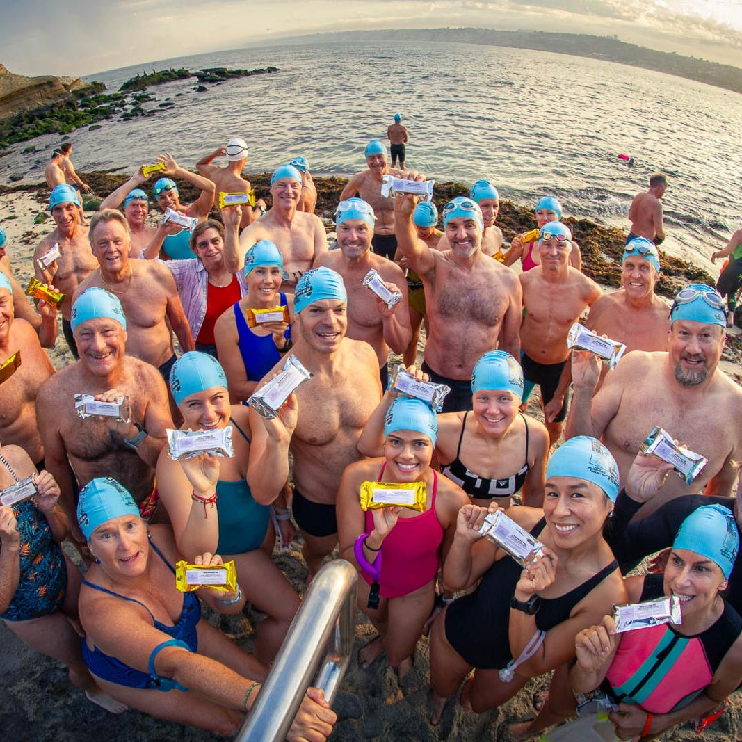 Group of swimmers on a beach at La Jolla Cove holding Shark Bars and wearing blue swim caps with ocean and sunrise in the background