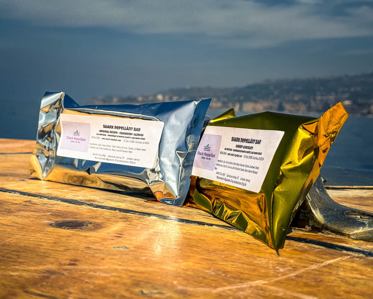 Two Shark Bars in shiny silver and gold foil packaging with ingredients labels on a wooden surface by a waterfront in southern california