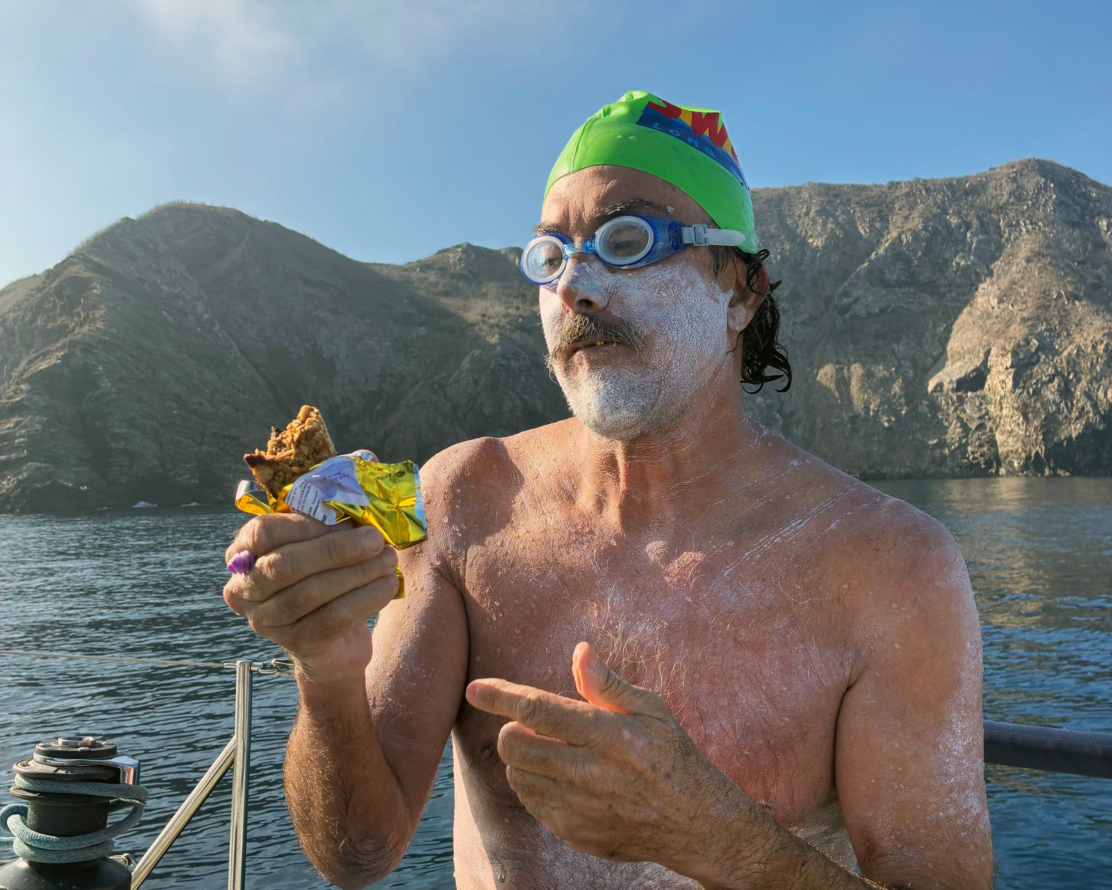 Channel swimmer with a mustache and white zinc sunscreen on face wearing swim cap and goggle eating a bite of a cherry chocolate oatmeal honey shark bar in front of catalina island while sitting on a boat in the ocean water